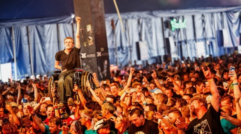 Groep mensen voor een festivalpodium. Een van de bezoekers wordt met rolstoel boven de menigte uit getild.