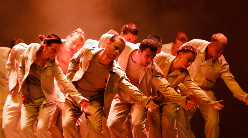 Een compacte groep van 12 dansers reikt synchroon omlaag met 1 arm. Ze zijn gekleed in diverse tinten wit en kaki.