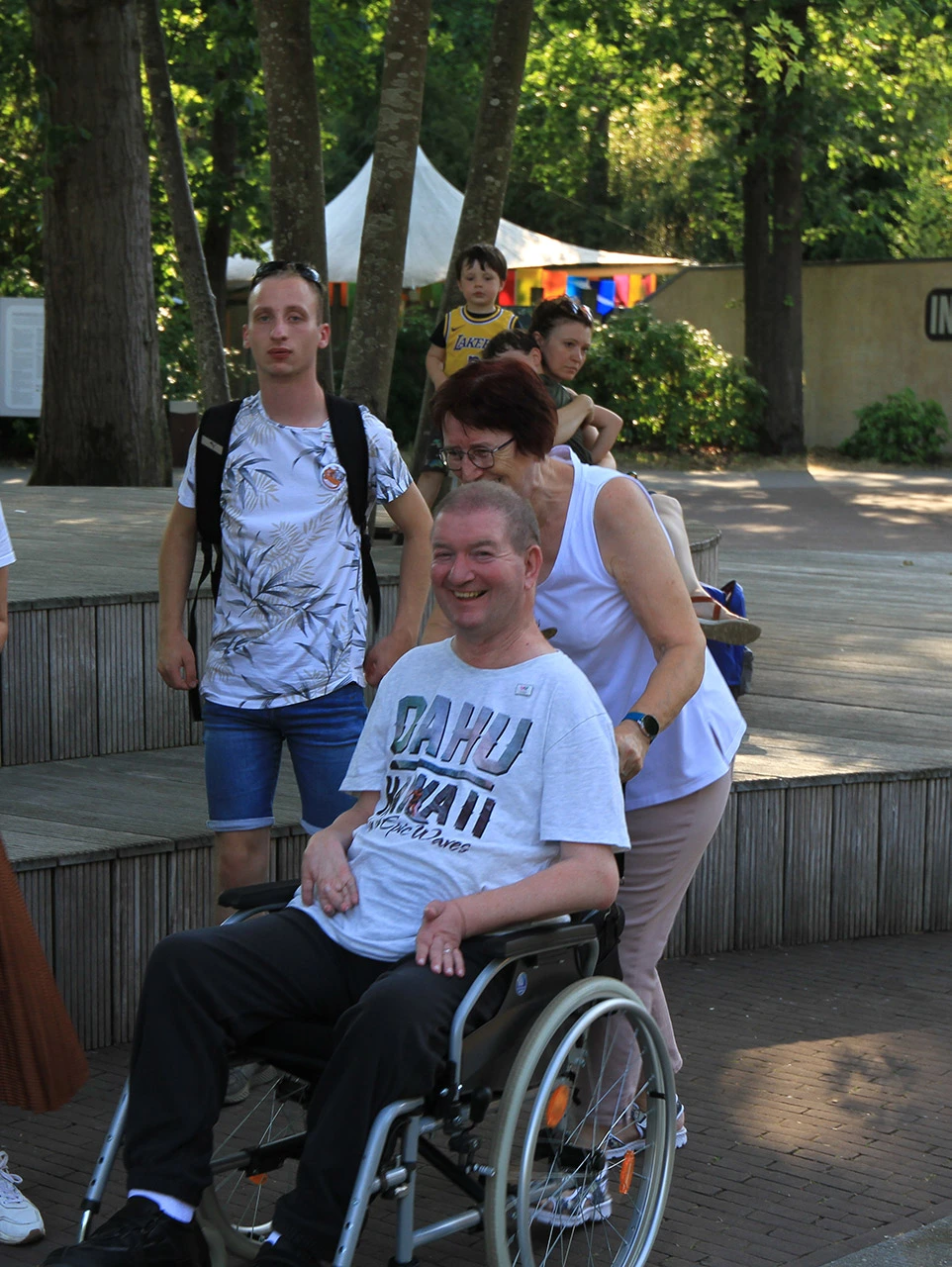 Een man van 65+ met heel kort haar en een zwarte joggingbroek aan met een wit t-shirt met tekst erop zit in een rolstoel. Hij lacht.