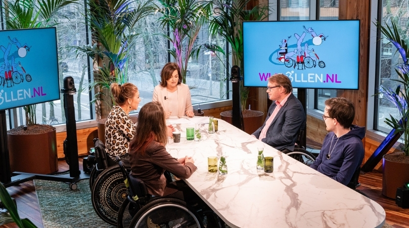 drie gasten in een rolstoel zijn in gesprek met een presentatrice aan een gesprekstafel, op de achtergrond staat een scherm met wijrollen.nl erop